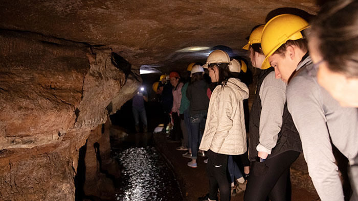Students on a field trip, underground, in a local cave. They are wearing hard hats and using flashlights to see the cave features.