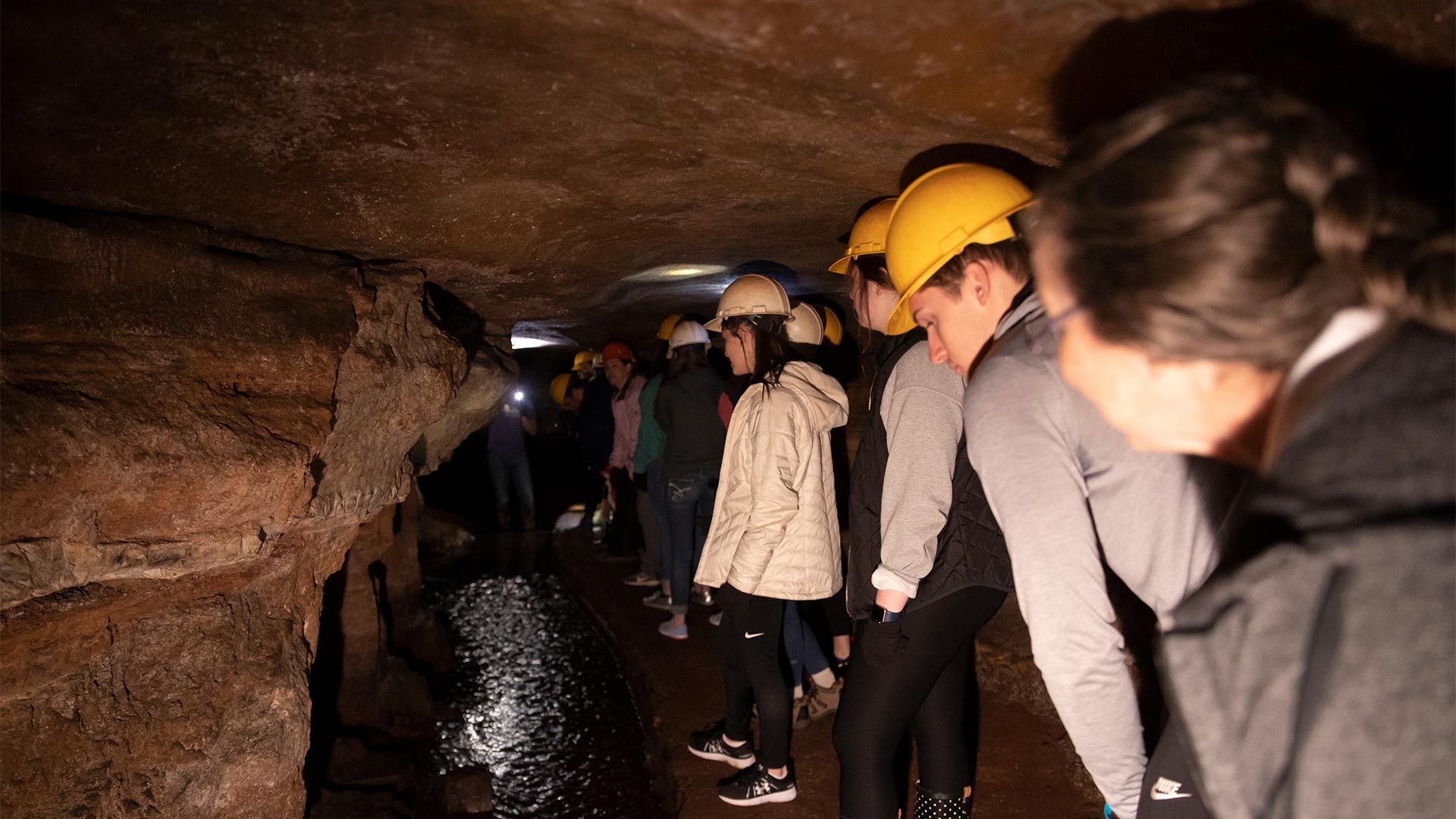 Students on a field trip, underground, in a local cave. They are wearing hard hats and using flashlights to see the cave features.