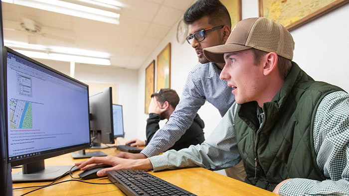 Student assisting another student with something on a computer monitor.