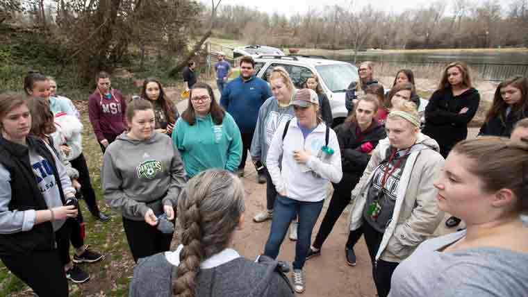 Group of students outdoors, listening, and gathered together around a faculty member.