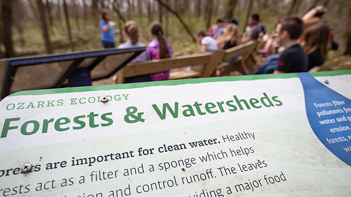 Partial Ozarks Ecology informational sign with a student lecture taking place in the background.